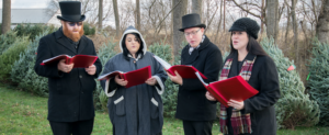 Four Christmas carolers singing next to a bunch of Christmas trees.