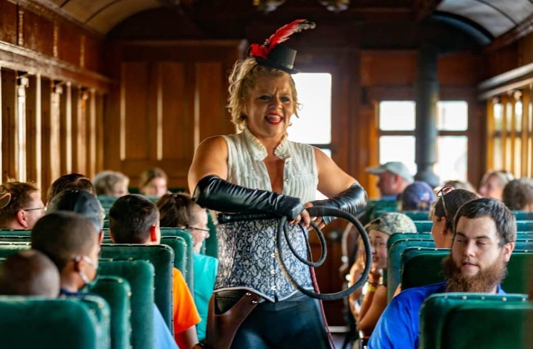 A group of people on a train watching a live performance of a lady in costume.