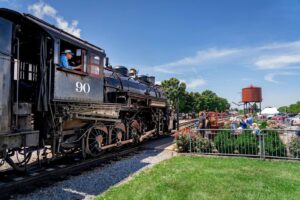 A steam train rolling along the tracks at Strasburg Rail Road while people on the sidelines wave and take pictures.