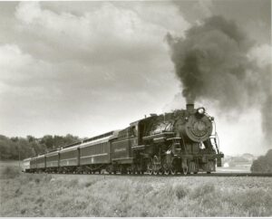 A black and white photo of a Vintage steam locomotive with smoke, traveling through countryside.