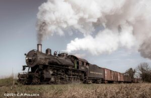 Steam locomotive emitting smoke travels through grassy field in beautiful railroad photography shot.