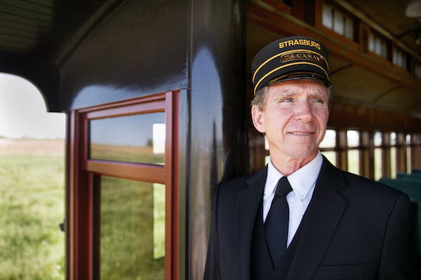 A smiling train conductor wearing his full, modern railroad conductor uniform on a Strasburg Rail Road train.