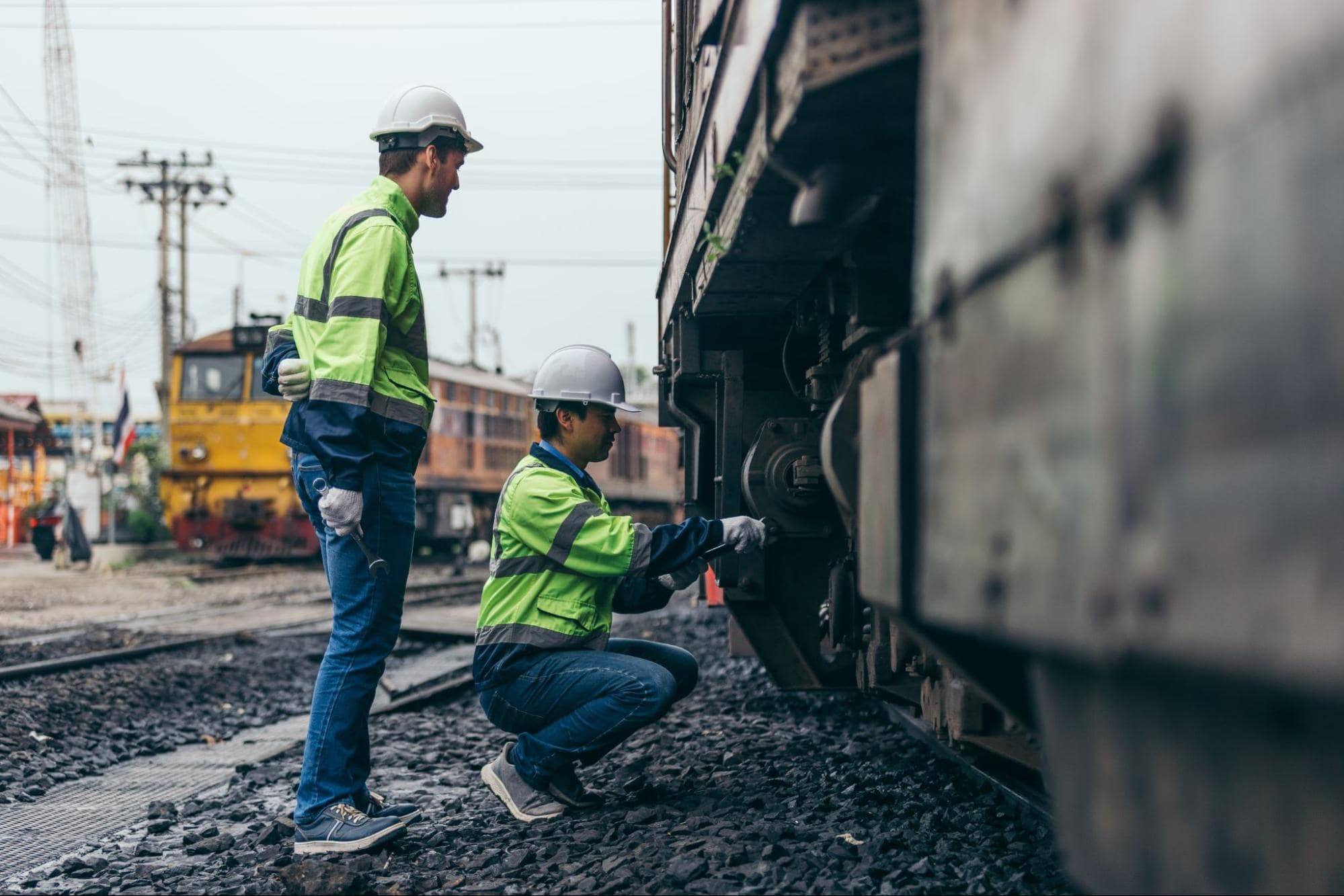 Two workers inspecting a train's wheels, both wearing high-visibility clothing for railroad workers, including hard hats and vests.