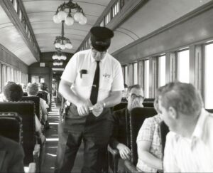 Black and white photo of a conductor checking tickets on a train, wearing a short-sleeved shirt and railroad uniform hat.